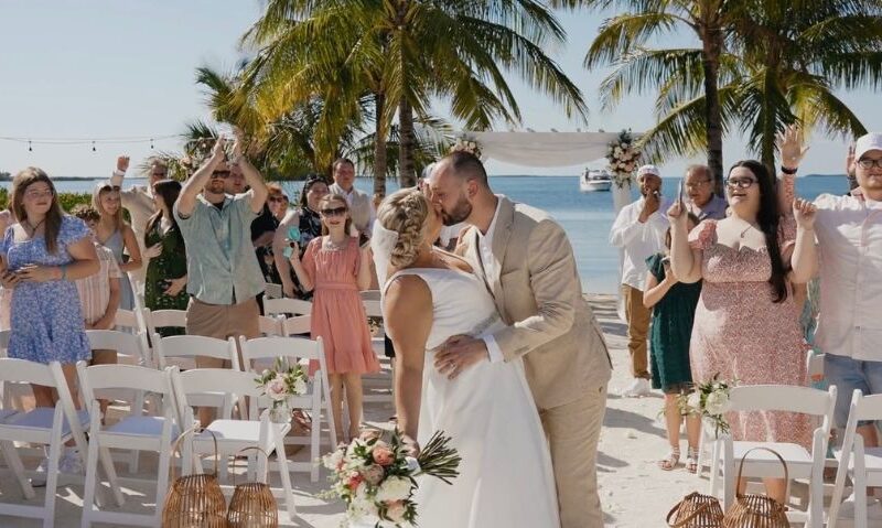 Couple kissing at the end of their beach wedding ceremony, surrounded by cheering guests under palm trees and string lights.