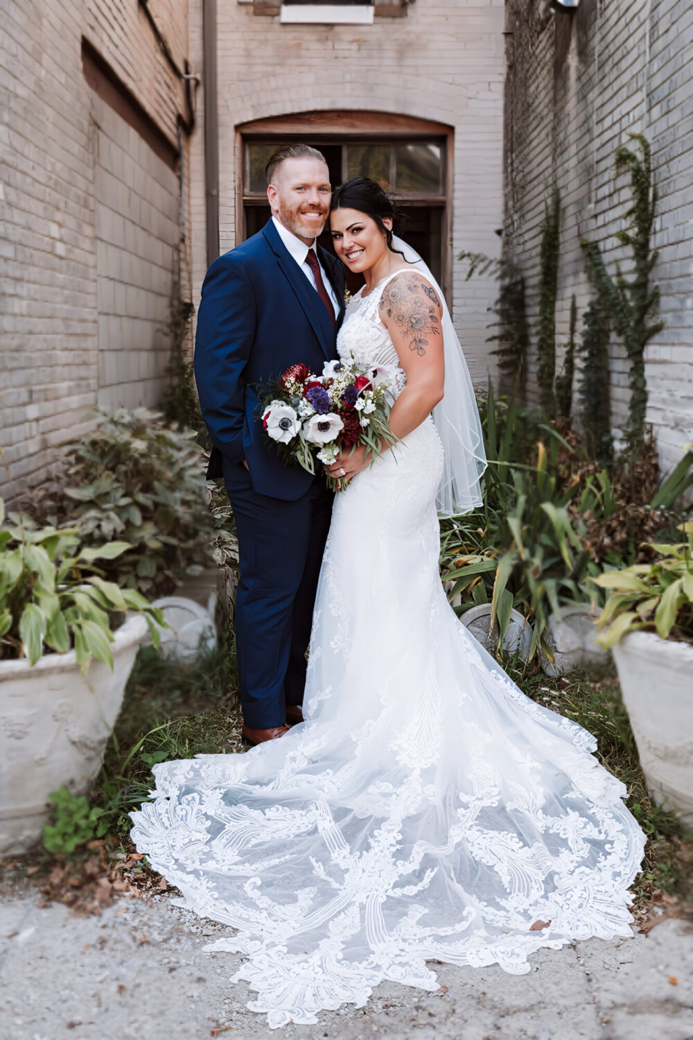 Bride in a detailed lace gown standing with her groom in a blue suit in a rustic alleyway, holding a bold bouquet of white and burgundy flowers.