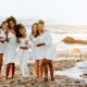 A group of seven women dressed in white pose together on a sandy beach at sunset, holding glasses of rosé and smiling.