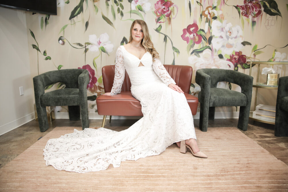 A bride lounges on a pink velvet couch wearing a long-sleeved lace wedding dress with a sweetheart neckline and a small front cutout.