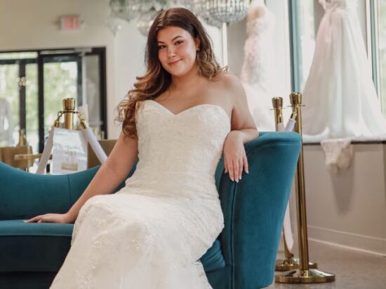 A bride in a fitted, strapless wedding gown with lace detailing poses on a teal couch in a bright bridal boutique.