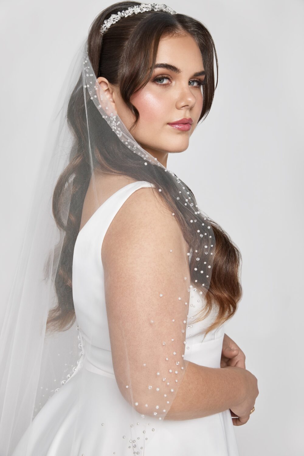 A bride stands in profile wearing a pearl-scattered fingertip veil with a crystal headpiece and classic gown.