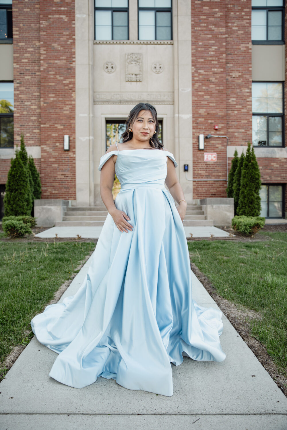A woman wears a soft blue off-the-shoulder A-line gown with pleating, posing in front of a brick and stone historic building.