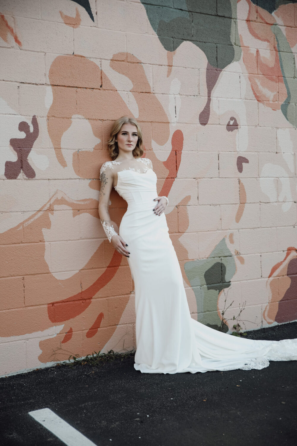 A bride leans against a floral mural wall wearing a sleek fitted gown with sheer lace sleeves and a bateau neckline with embroidered detailing.