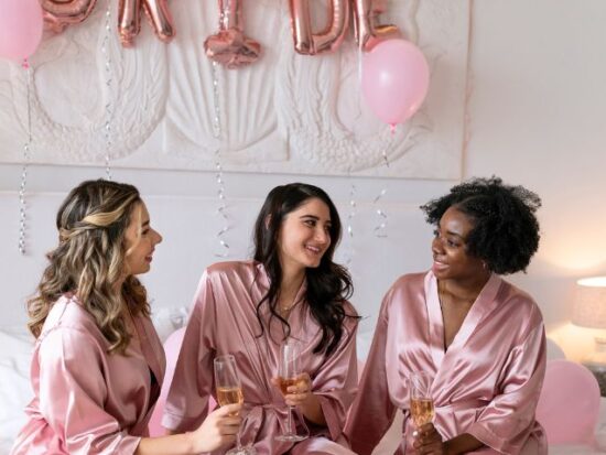Three bridesmaids in matching pink satin robes toast with champagne while sitting cross-legged on a bed decorated with pink balloons and soft lighting.