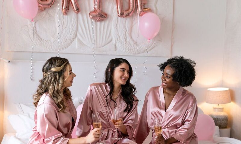 Three bridesmaids in matching pink satin robes toast with champagne while sitting cross-legged on a bed decorated with pink balloons and soft lighting.