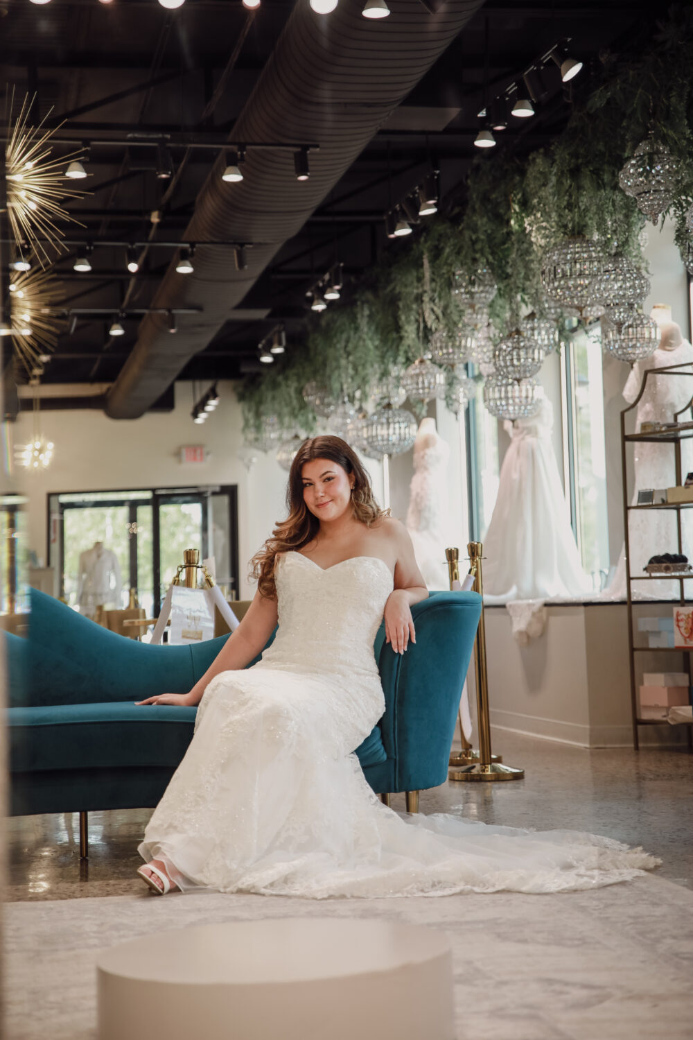 Bride in a strapless lace wedding dress sits confidently on a blue couch inside a modern boutique filled with dresses.