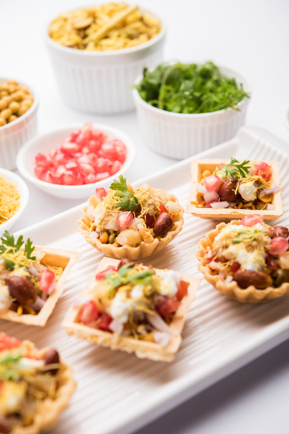 Mini taco bites on a white tray with bowls of toppings like pomegranate seeds, cilantro, and chickpeas.