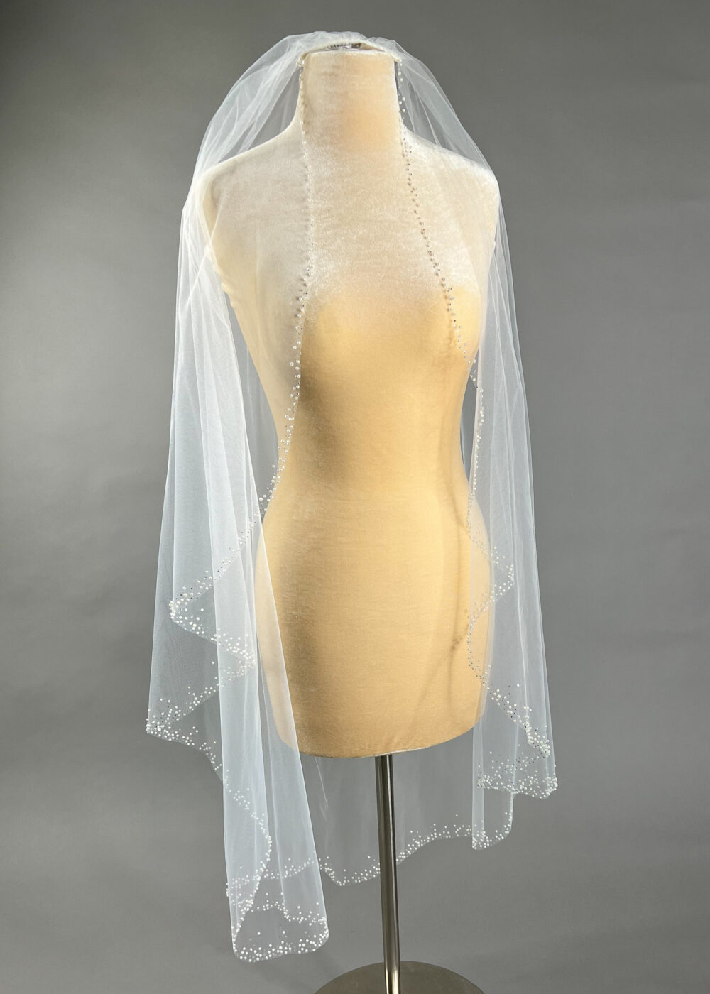 A fingertip-length wedding veil with pearl edging is displayed on a mannequin against a gray backdrop.