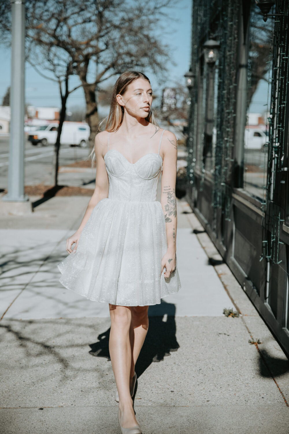 A woman in a short, glittering wedding reception dress walks outdoors on a sunny day with tattoos visible on her arm.