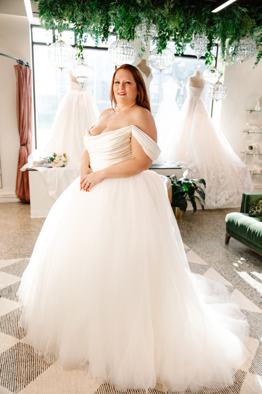 Bride in an off-the-shoulder gown with full tulle skirt poses in a softly lit showroom surrounded by gowns on mannequins.