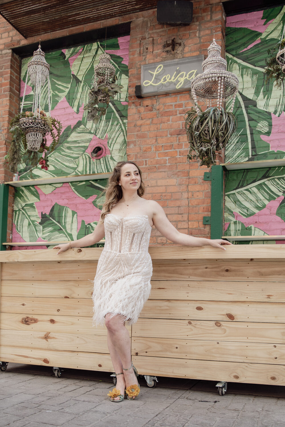 A bride poses in a strapless beaded mini dress with feathered skirt in front of a tropical mural backdrop.