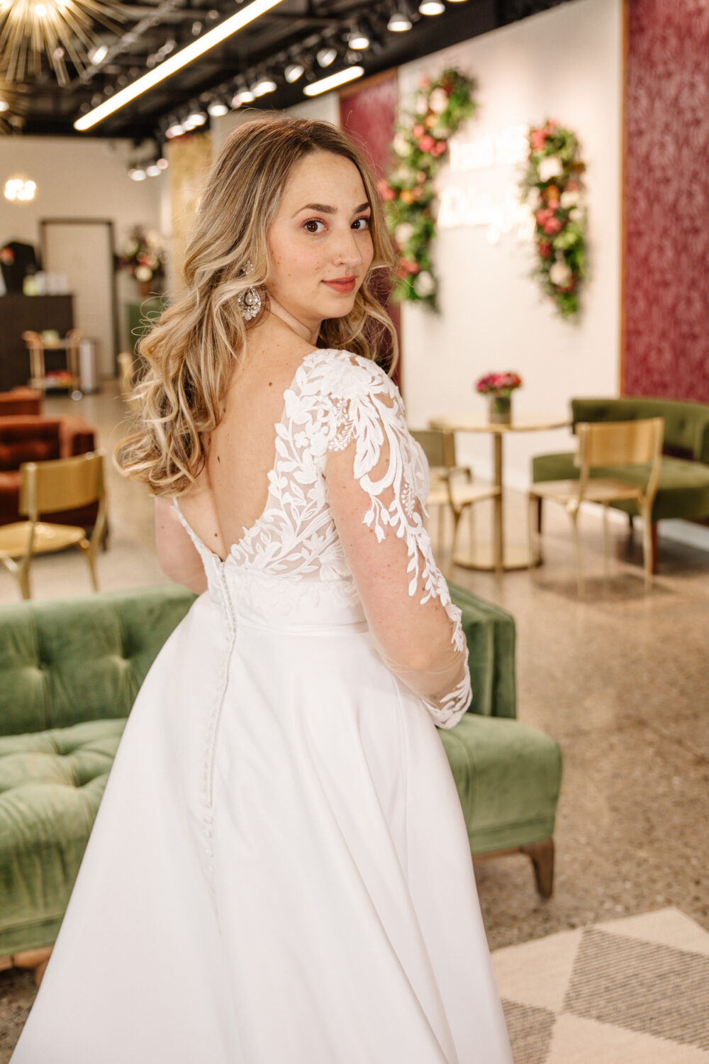 A bride in a long-sleeve lace backless gown poses gracefully inside a bridal boutique, glancing back with soft curls cascading over her shoulder.