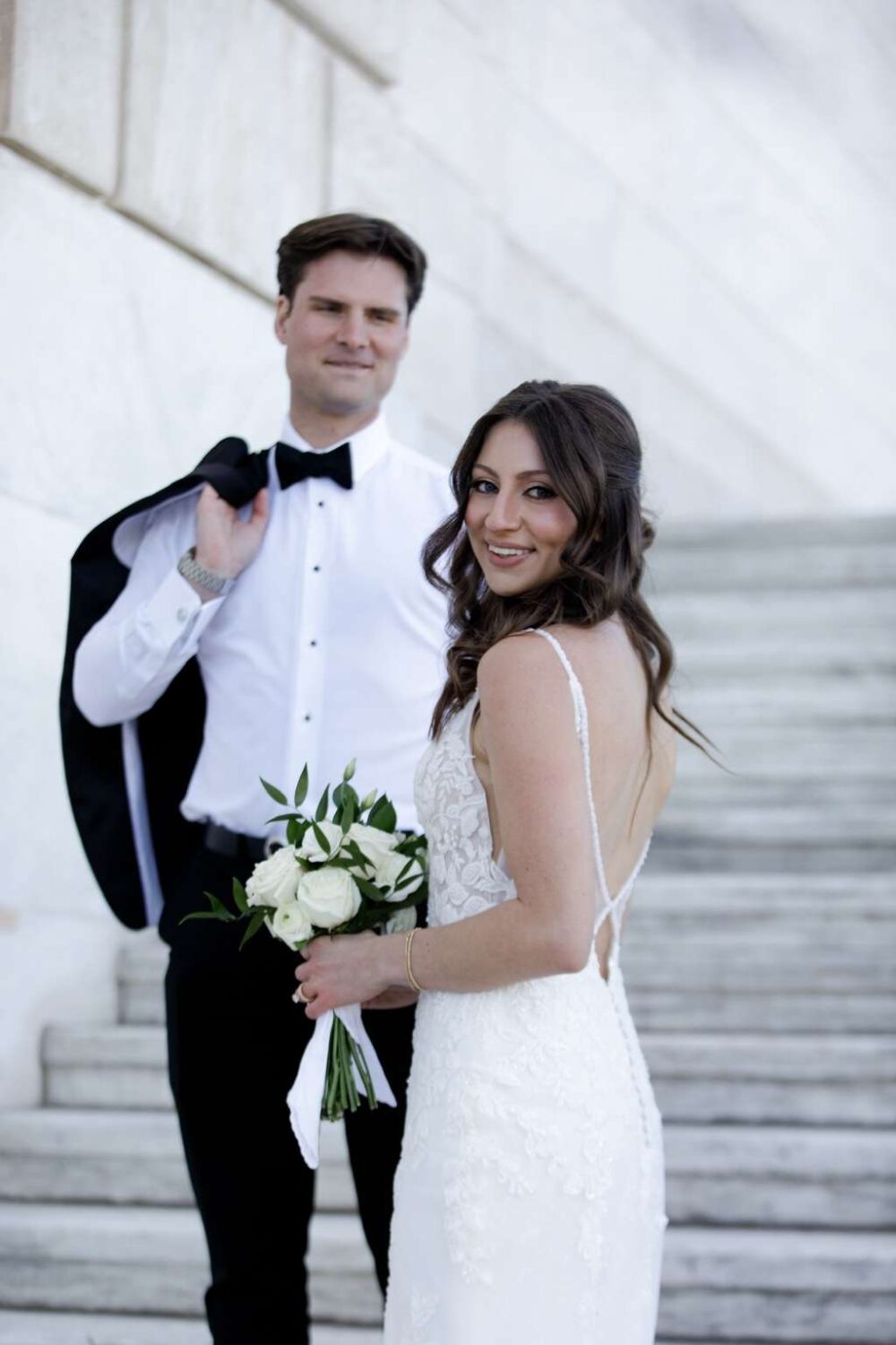 A bride in a fitted lace backless wedding dress stands with her groom on marble steps, holding a bouquet of white roses and smiling at the camera.