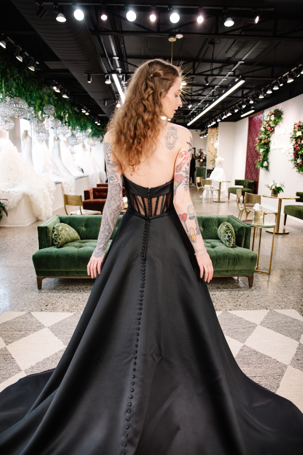 A bride with long curly hair and tattoos models a strapless black backless wedding dress with a dramatic train in an elegant bridal boutique.