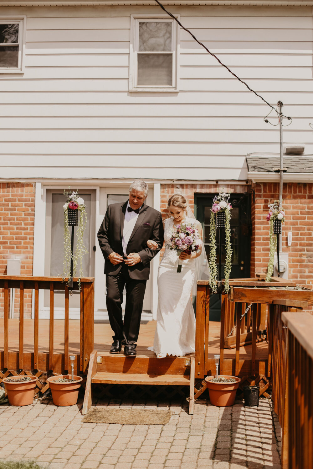 A bride walks arm-in-arm with her father down the wooden steps of a backyard deck decorated with hanging florals and greenery.
