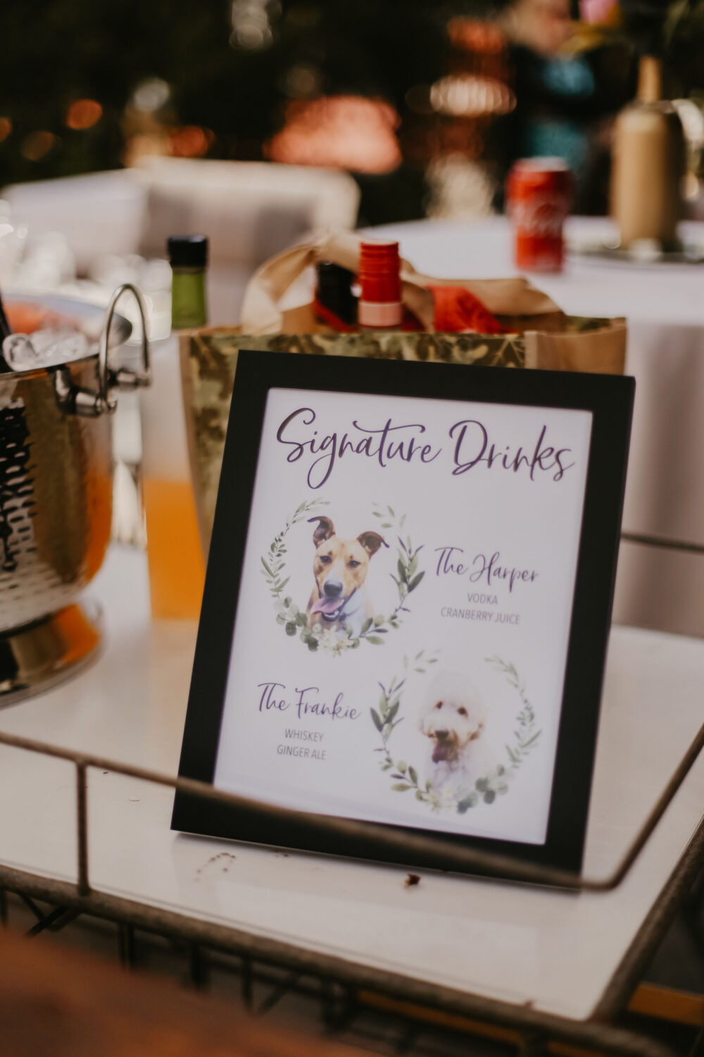 A framed wedding sign on a drink table displays “Signature Drinks” featuring custom cocktails named after two dogs, with illustrated portraits of each.