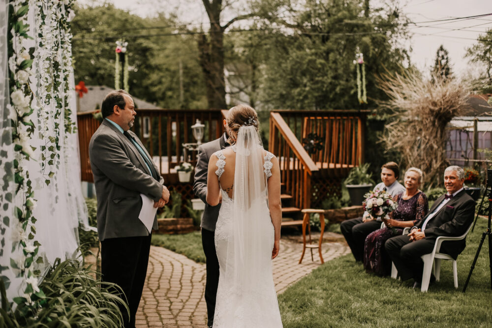 The bride stands facing the officiant and groom at the altar during a backyard ceremony, with seated guests including family members watching nearby.