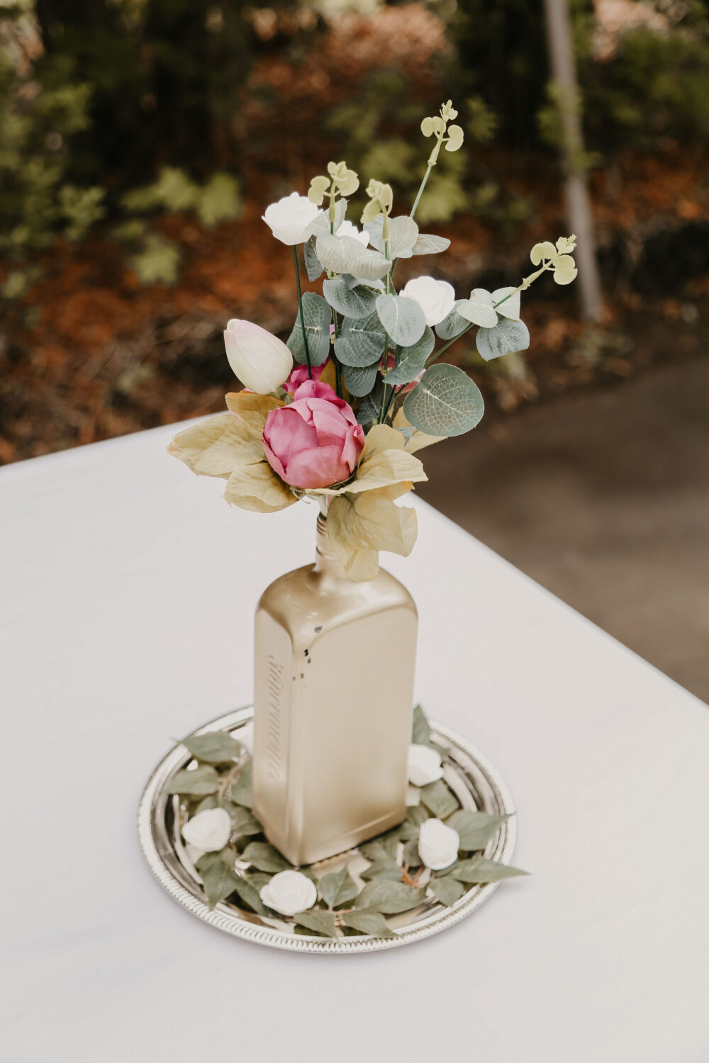 A painted gold bottle serves as a vase for a simple wedding table centerpiece, filled with artificial flowers and eucalyptus, placed on a silver tray.