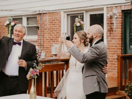 The bride, groom, and a man in a tuxedo raise their glasses in a toast on a wooden backyard deck, surrounded by floral wedding decorations.