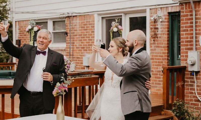 The bride, groom, and a man in a tuxedo raise their glasses in a toast on a wooden backyard deck, surrounded by floral wedding decorations.
