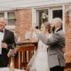 The bride, groom, and a man in a tuxedo raise their glasses in a toast on a wooden backyard deck, surrounded by floral wedding decorations.