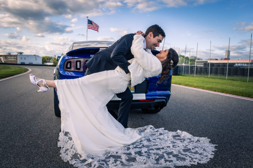 Groom dips smiling bride in front of a blue muscle car on a racetrack with an American flag in the background.