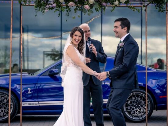 Bride and groom laughing during an outdoor wedding ceremony in front of a blue sports car.