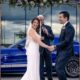 Bride and groom laughing during an outdoor wedding ceremony in front of a blue sports car.