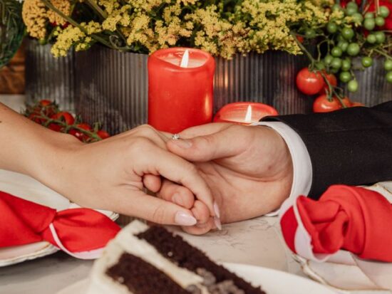 A close-up shot captures the bride and groom gently holding hands on the table next to a slice of chocolate cake and red candles.