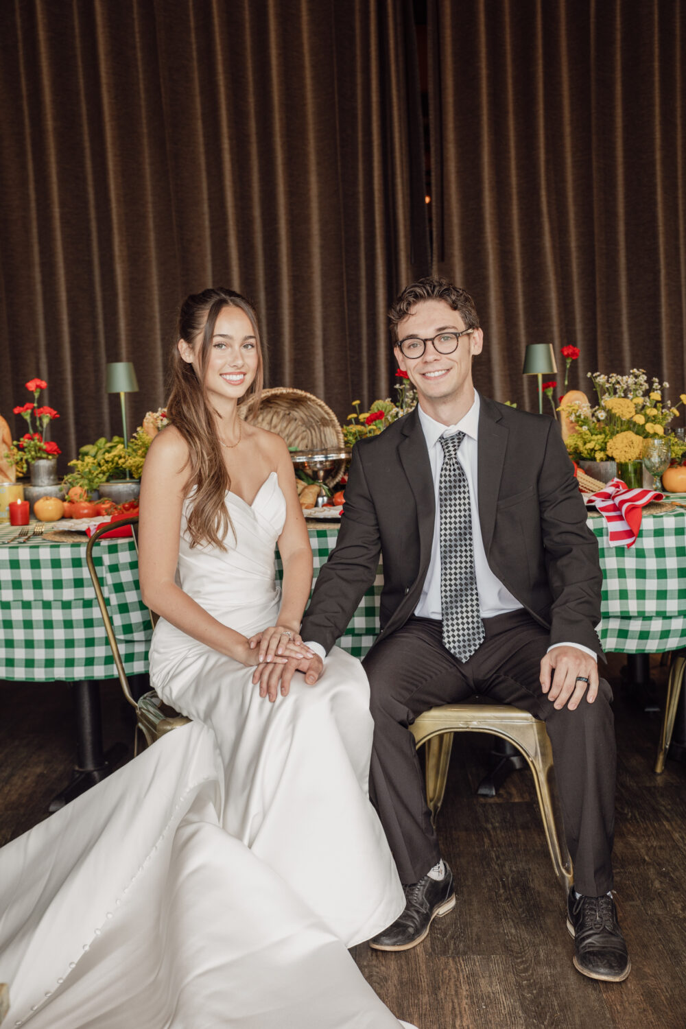 The newlyweds sit side by side smiling, hands intertwined, in front of a festive reception table decorated with flowers and checkered linens.