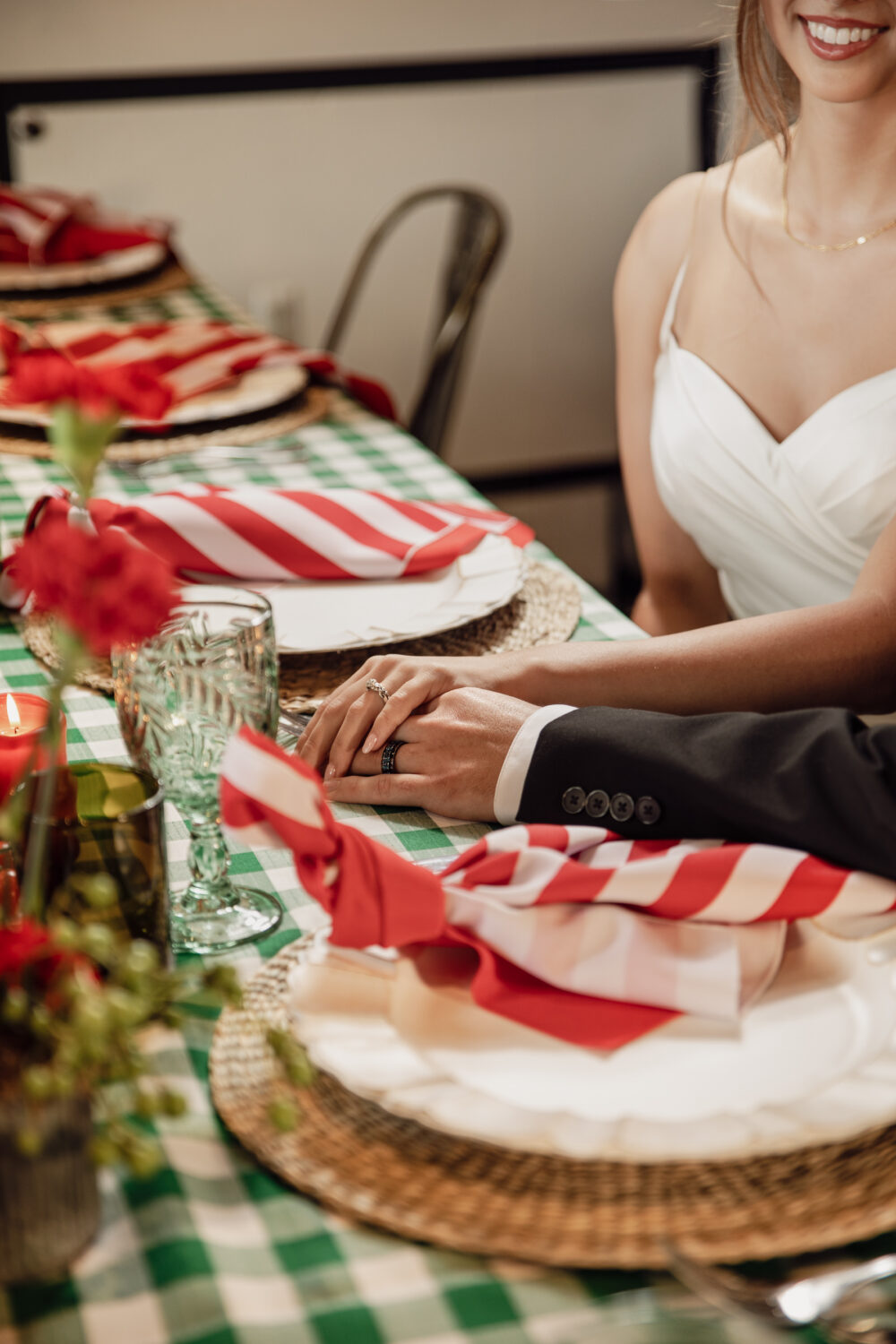 A bride and groom sit at a table with green and white checkered linens, holding hands over place settings with red and white striped napkins.