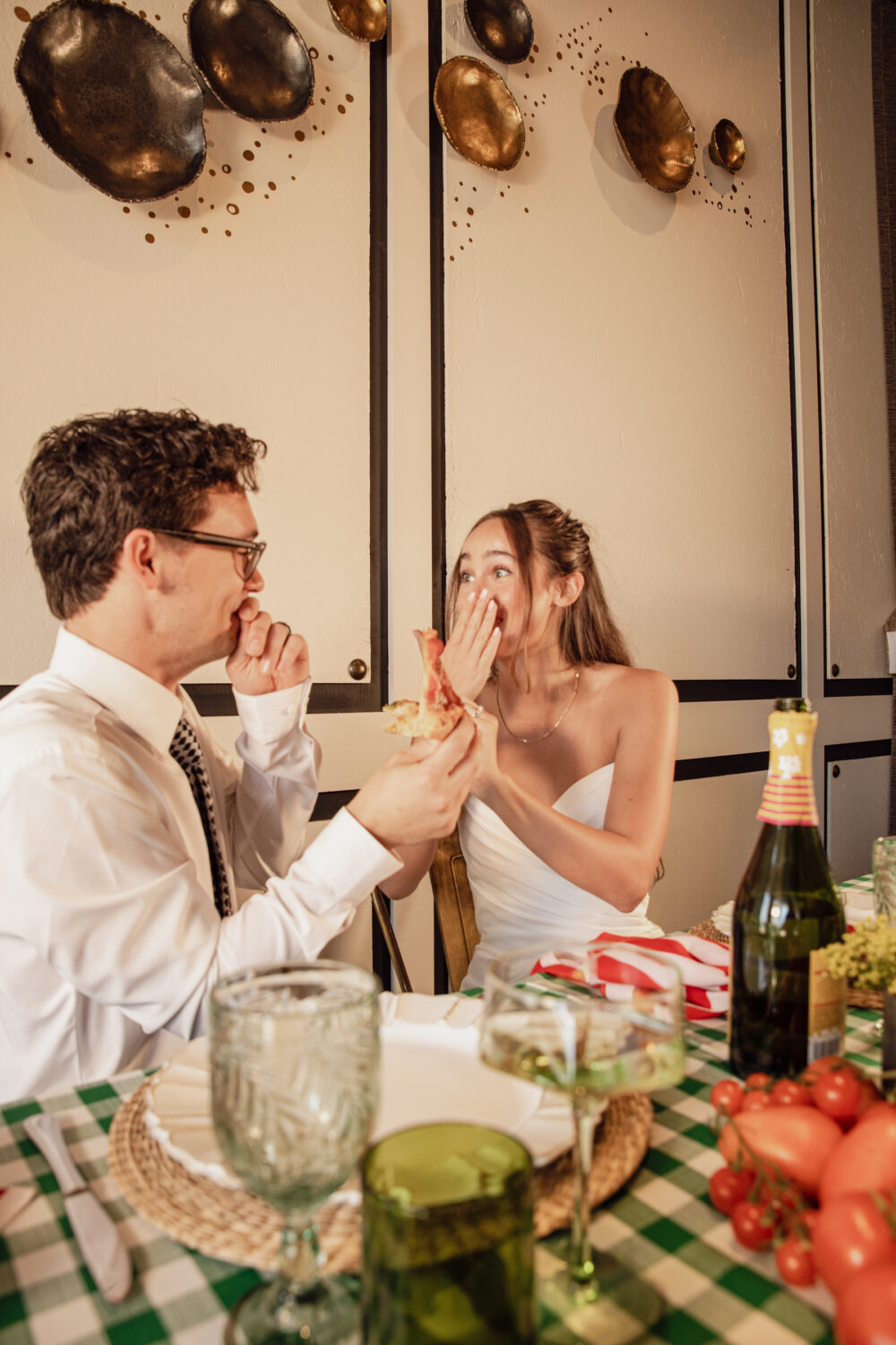 The bride and groom sit across from each other at a small table, sharing dessert and holding hands in front of a vertical garden with artistic wooden boards.