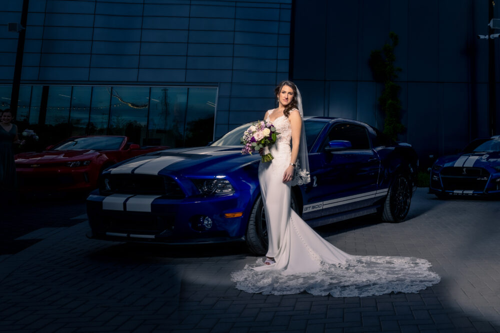 Bride standing confidently in front of a row of sports cars outside a sleek venue building.