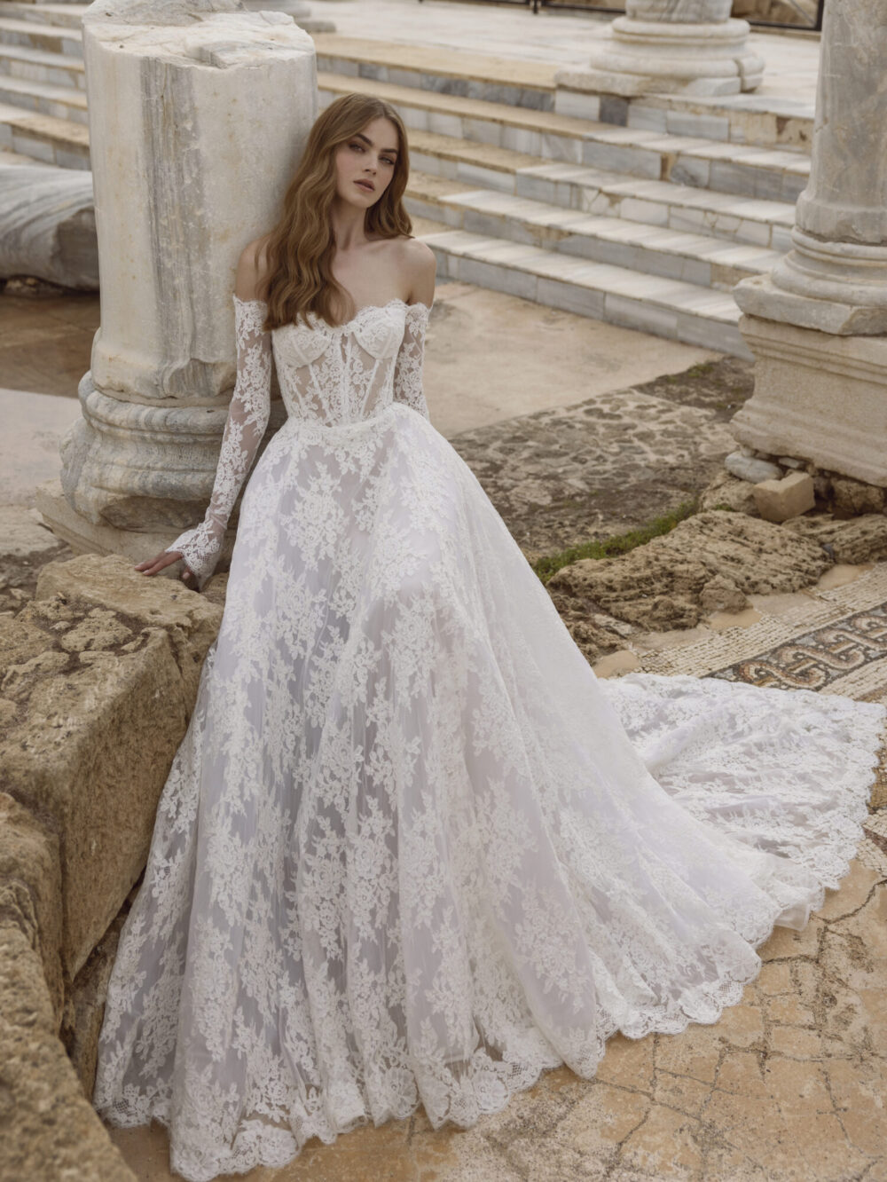 A model wearing a white lace off-the-shoulder wedding dress in an Athens-themed location, leaning towards a ruined pillar.