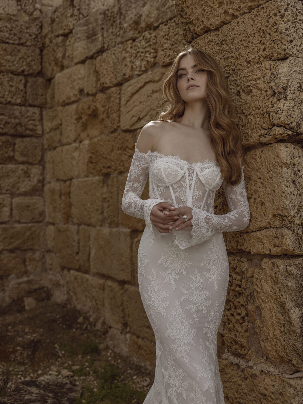 A model wearing a white lace off-the-shoulder trumpet wedding dress, leaning on stone brick walls.