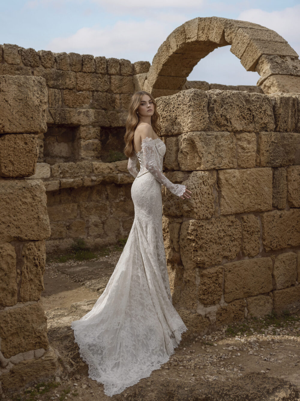 A model wearing a white lace off-the-shoulder trumpet wedding dress in a location with large stone brick walls, showing the back details of the dress.