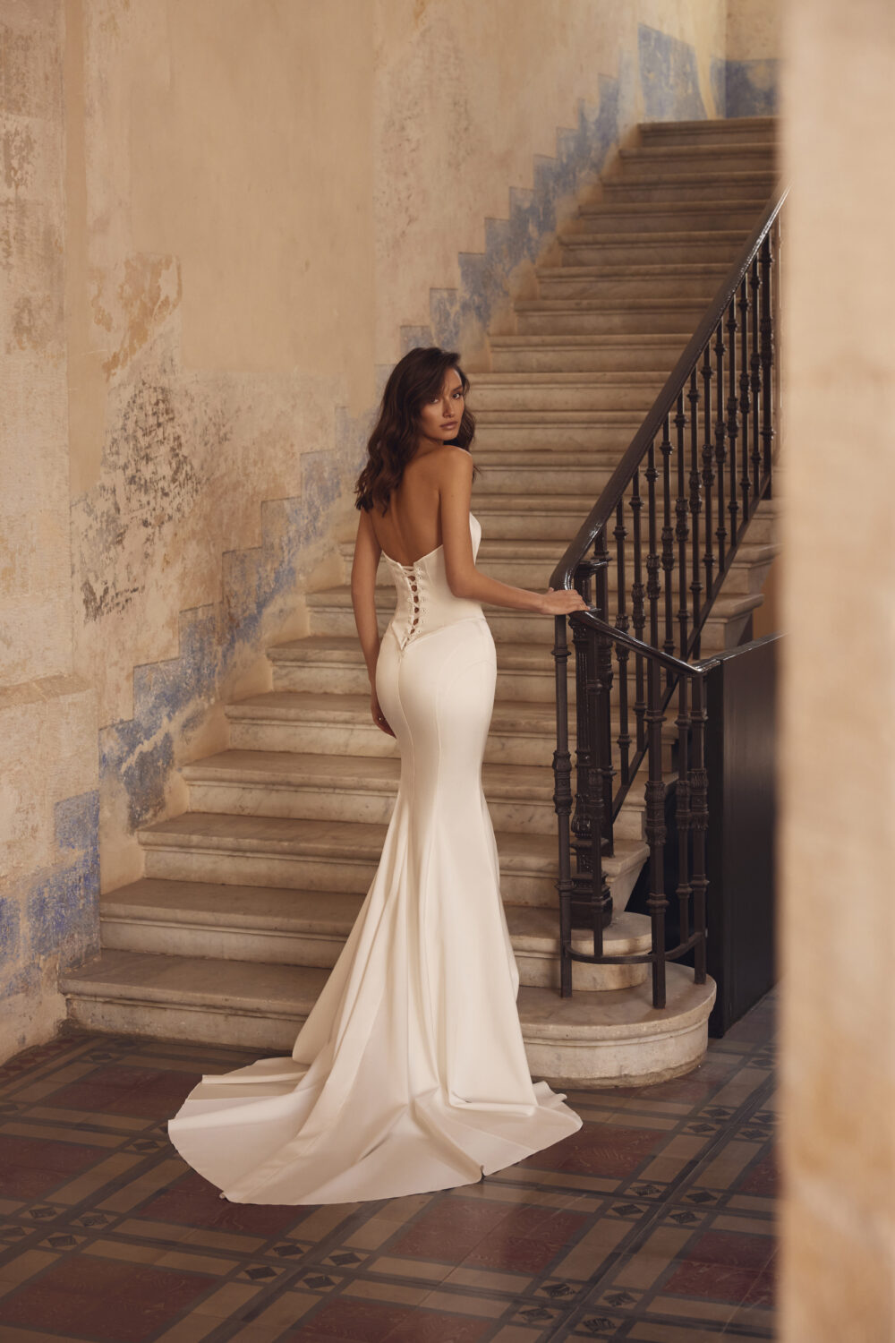 A brunette model wearing a satin white trumpet wedding dress by the staircase of an aged house.