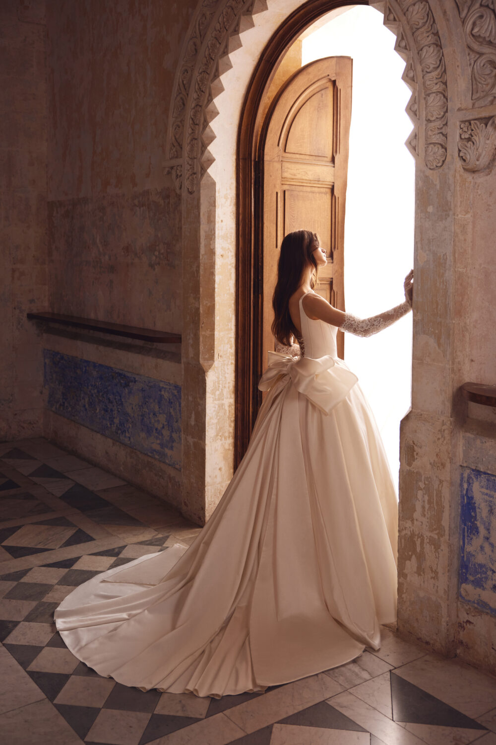 A Pnina Tornai satin ball gown with a large bow is worn by a model while looking outside from the door.