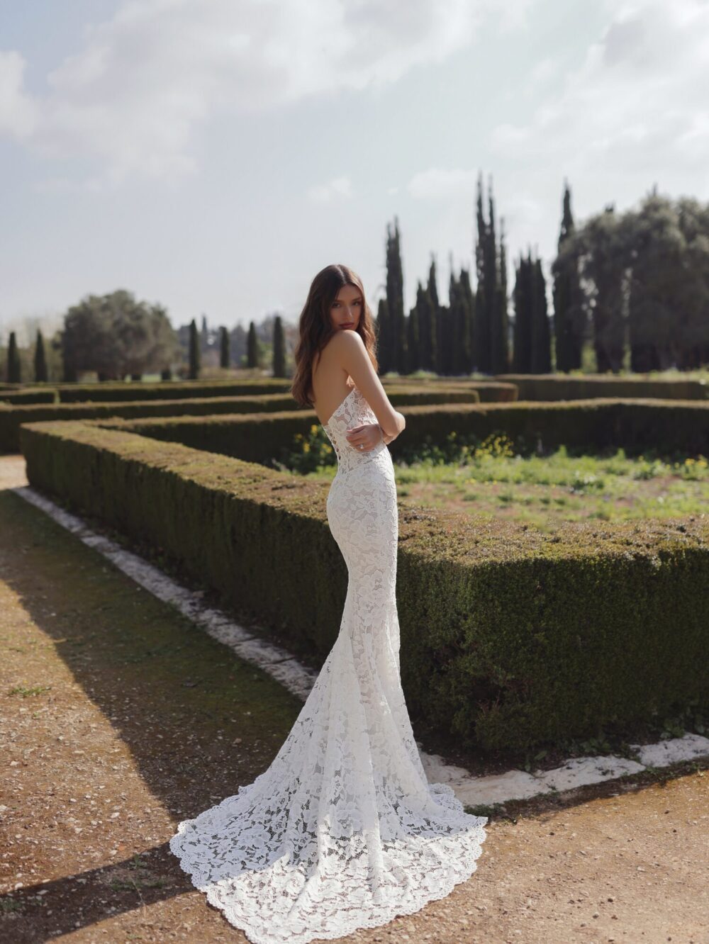 A bride wearing a lace tube wedding dress with a short train is embracing herself in a massive garden with a hedge maze and tall trees.