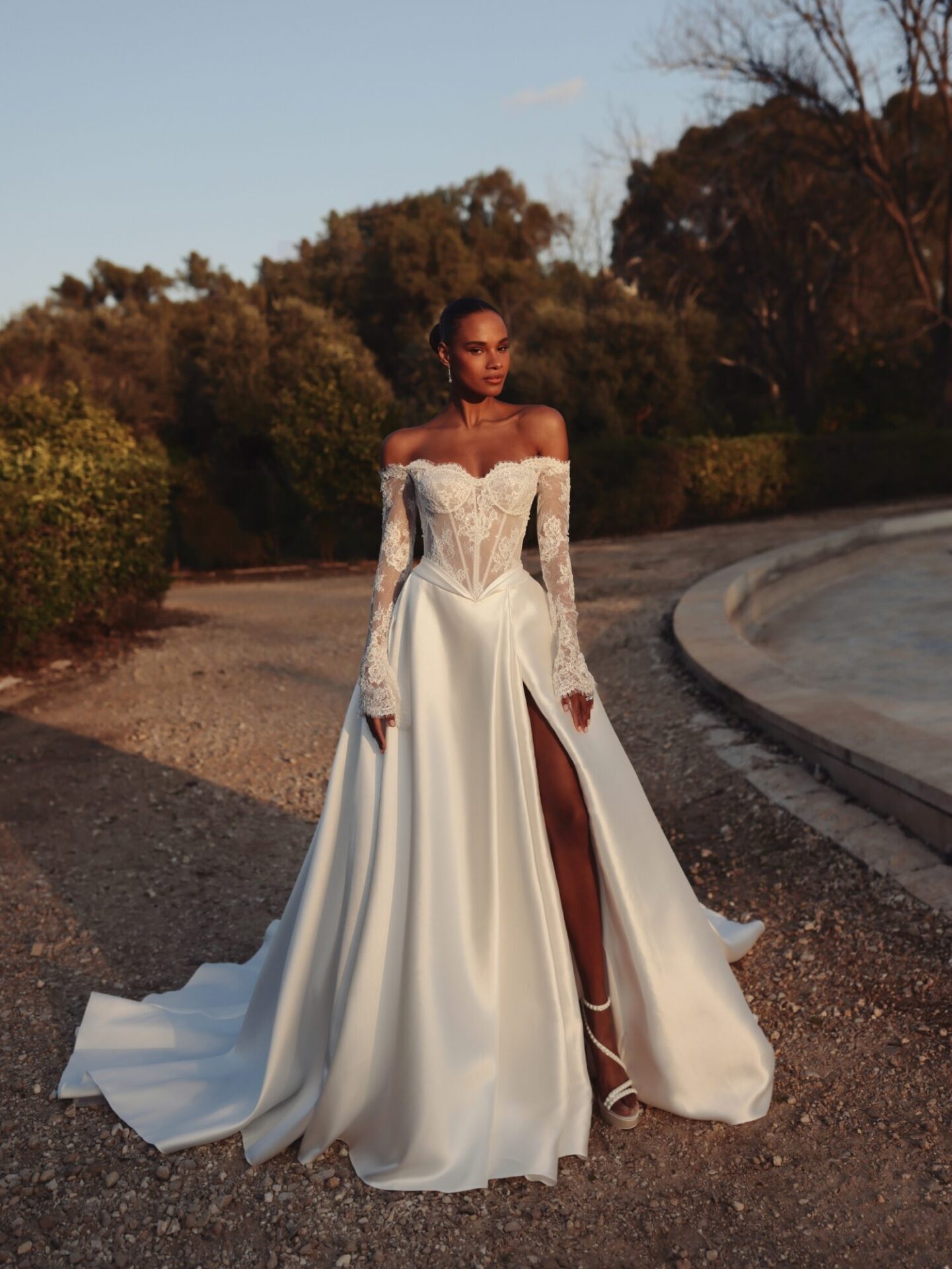 A model is wearing a off-the-shoulder lace top and satin bottom wedding dress with a thigh-high slit with her hair up and posing with one leg forward.