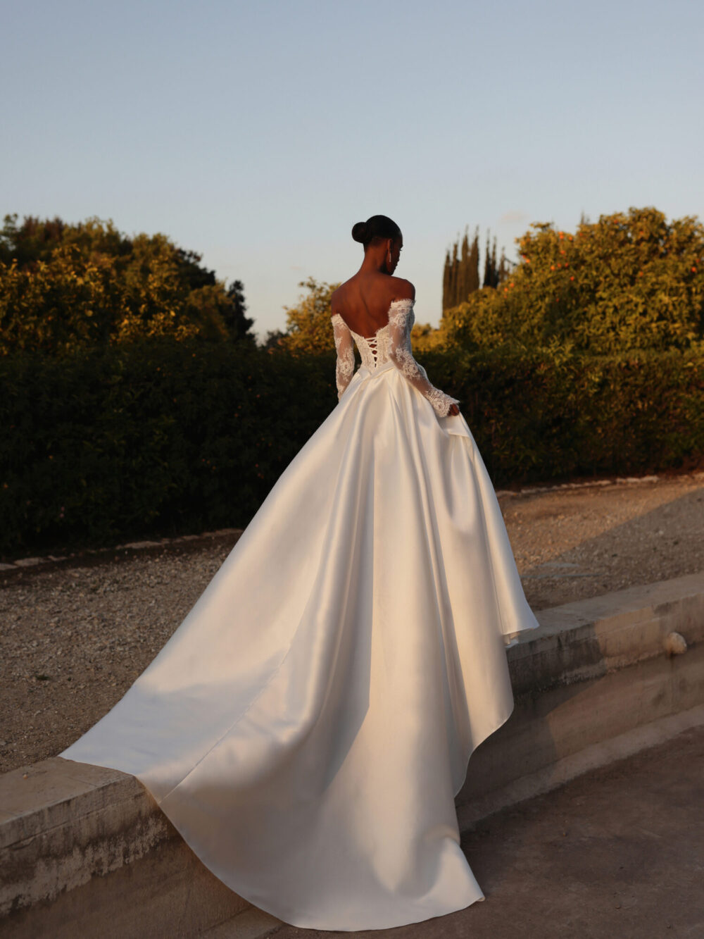 A model wearing a lace and satin wedding gown is walking along a driveway.
