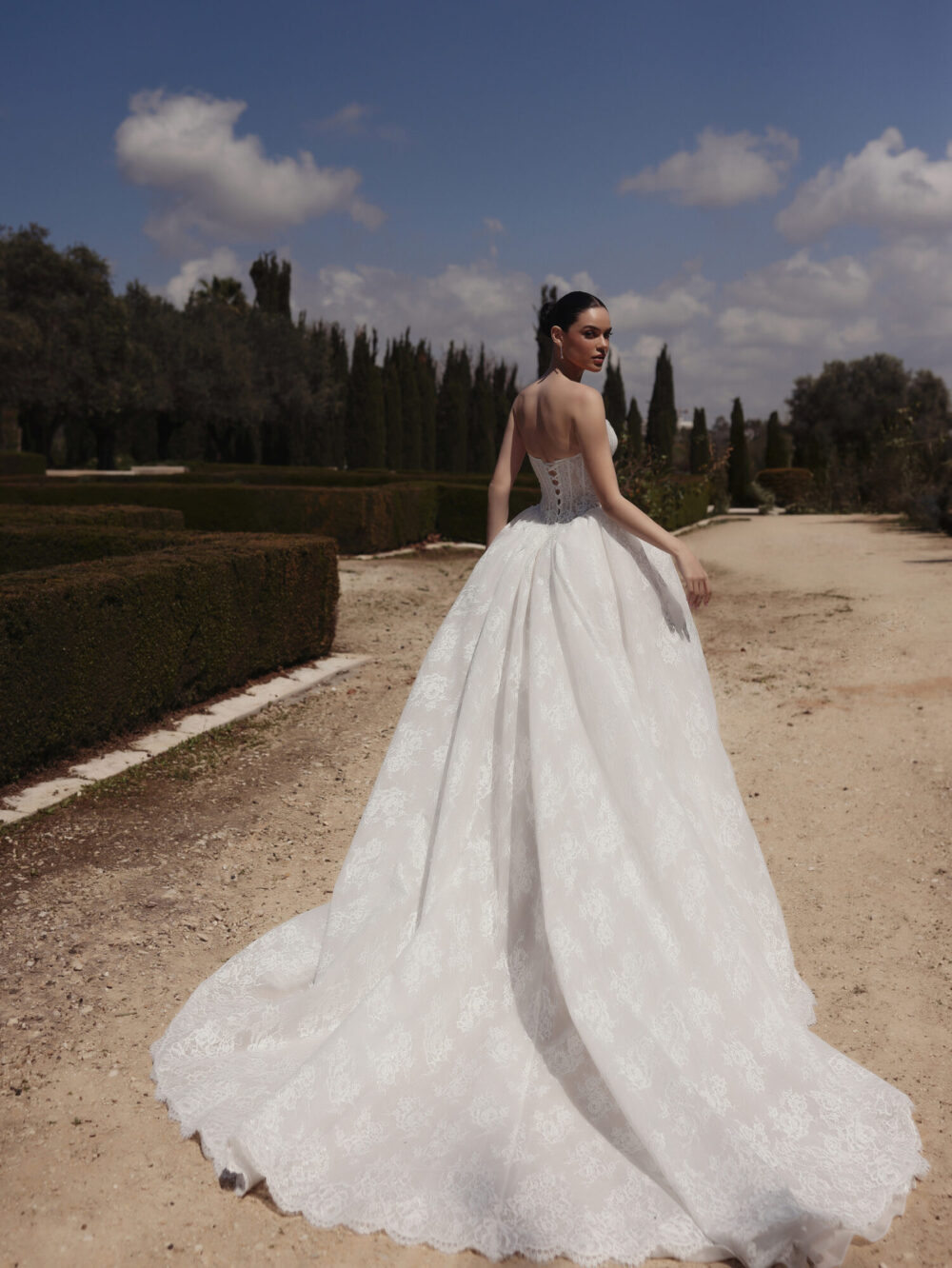 A lace ball gown is worn by a bride who is looking back while walking away in a garden.