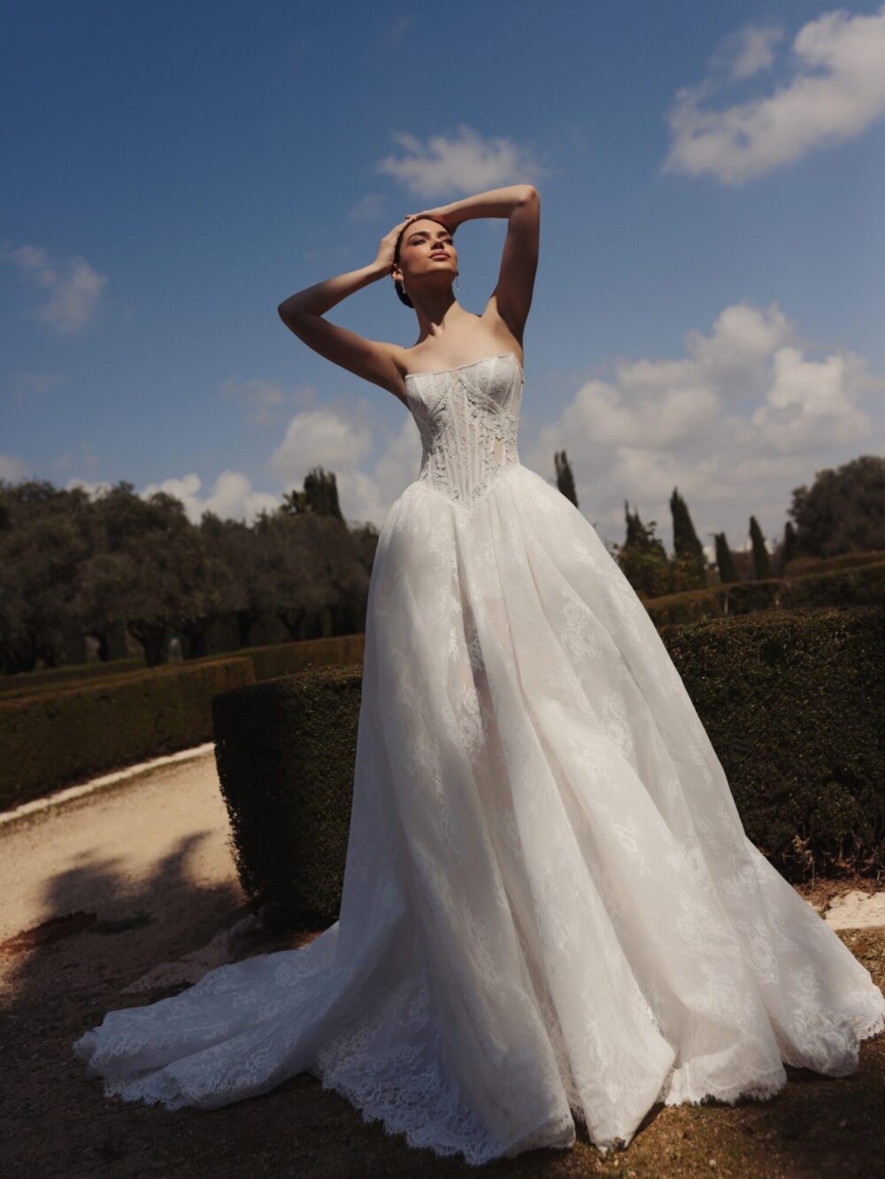 A lace ball gown with a ribbed tube bodice is worn by a model with both her hands on her head.
