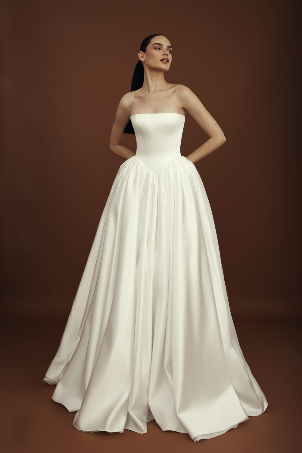 A model with long black hair is wearing a very timeless satin tube wedding dress with both her hands on her back.