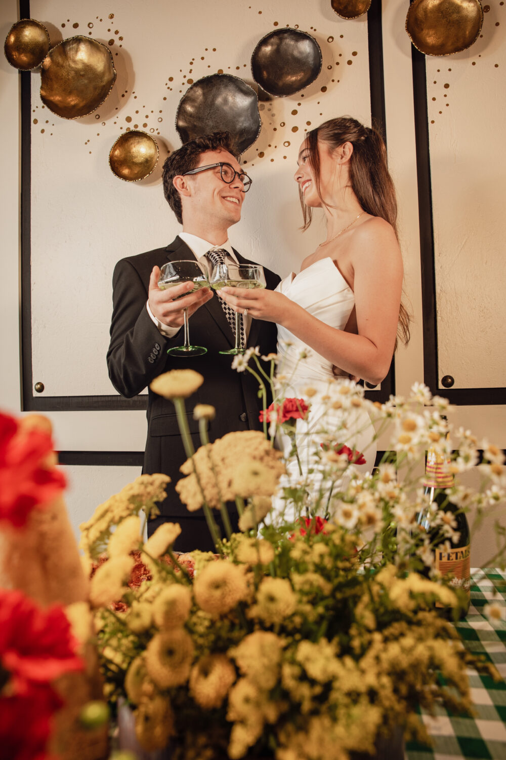 A smiling couple dressed in wedding attire clink glasses while standing in front of a modern wall with metallic decorative art.