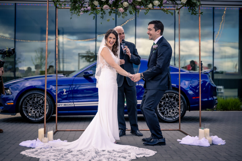 Bride and groom exchange vows outdoors under floral arch with blue GT500 Mustang behind them.
