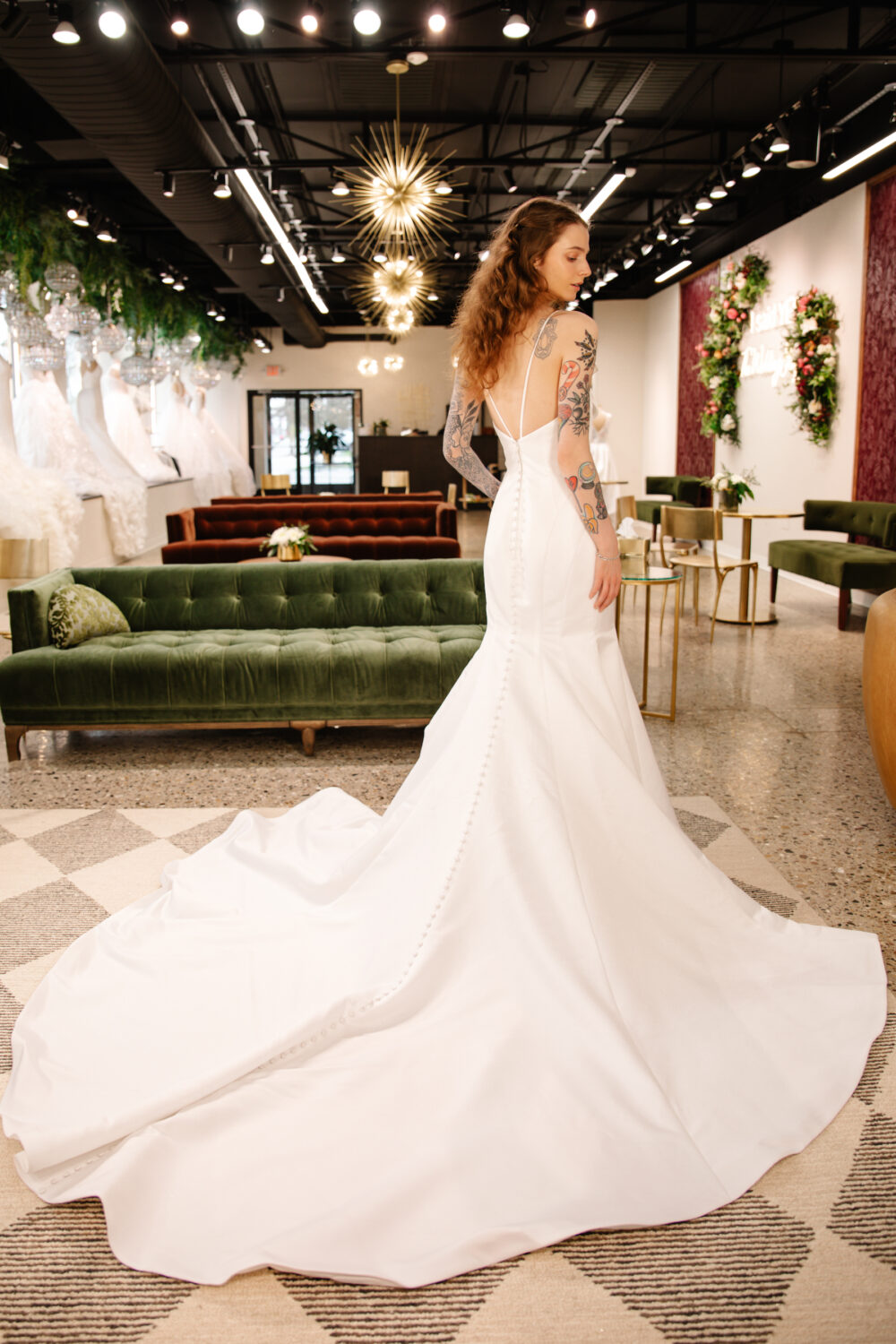 A tattooed bride in a fitted white backless gown with a long train poses elegantly in a well-lit bridal shop with green velvet sofas and floral decor.