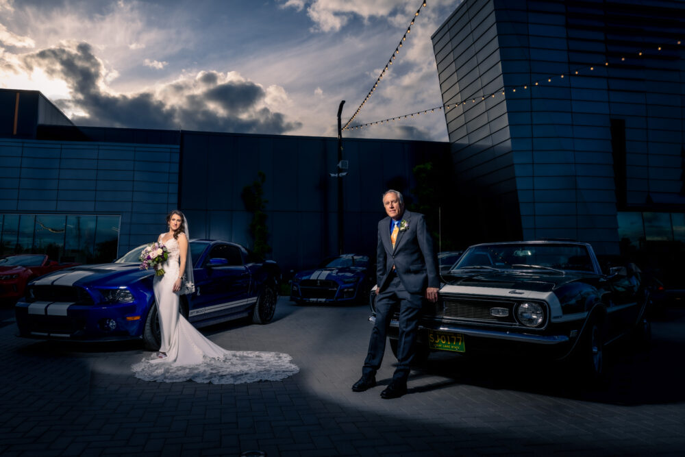 Bride and father posing with classic cars outside a modern building at sunset.