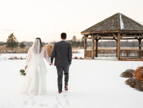 Bride and groom walking hand in hand through the snow toward a wooden gazebo on a winter day.